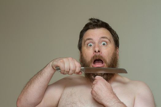 Man looks scared while cutting his own beard hair with a sharp knife