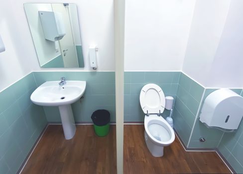 wide angle view of a toilet with the wooden floor and tiled wall with a mirror, a green basket, a sink and the ceramic bowl.