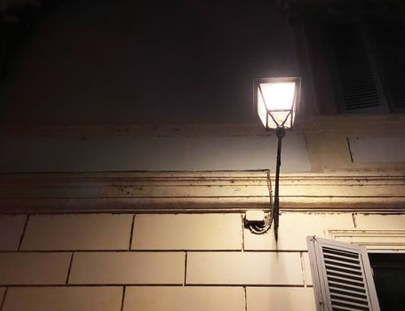 Antique decorated wrought-iron street lamp attached to the wall of an old building in the historic center of Rome at night. Glare of light on the wall. Night shot.