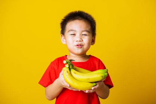 Happy portrait Asian child or kid cute little boy attractive smile wearing red t-shirt playing holds bananas comb fruit, studio shot isolated on yellow background