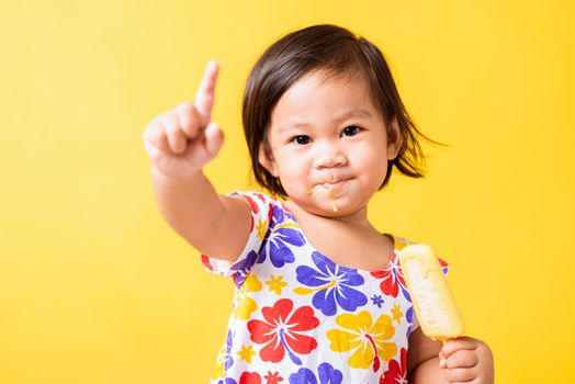 Happy portrait Asian baby or kid cute little girl attractive laugh smile wearing dick pattern shirt holds and eating sweet wooden ice cream, studio shot isolated on yellow background, summer concept