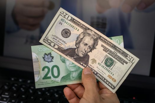 A hand holding two banknotes, one of twenty American dollars and the other of twenty Canadian dollars