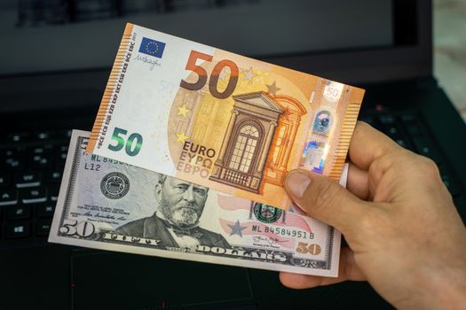 A hand holding two banknotes, one of fifty American dollars and the other of fifty euros