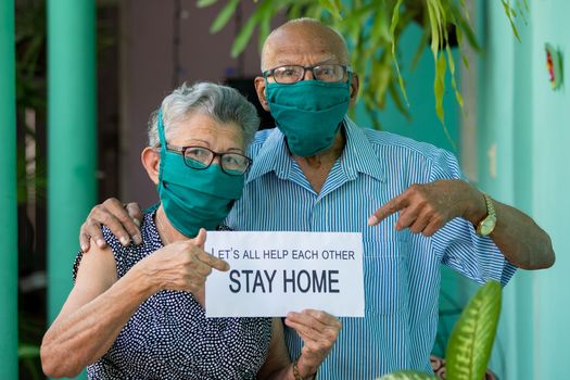 An elderly couple, both wearing face mask and glasses and holding an stay home sing