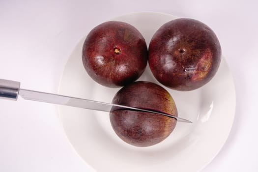 Three caimito fruits on a plate and white background, one of them cut in the middle by a knife.