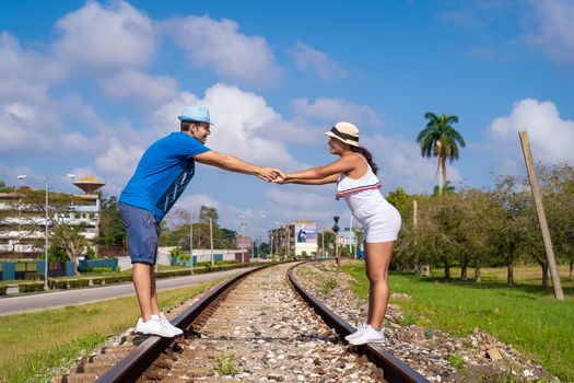 Couple of young people wearing hat held by their hands standing on train line, A sky with some clouds in the background