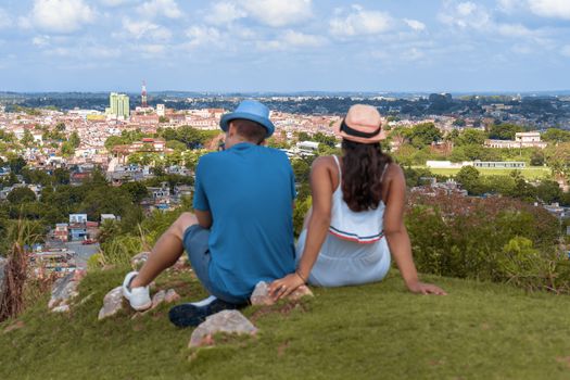A couple of young people wearing hat sitting on the floor looking towards a city from a hill
