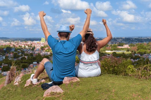 A couple of young people wearing hat with their arms up sitting on the floor looking towards a city from a hill