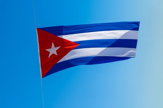 A Cuban flag with three blue an two white stripes, and a red triangle with a white star. Blue background