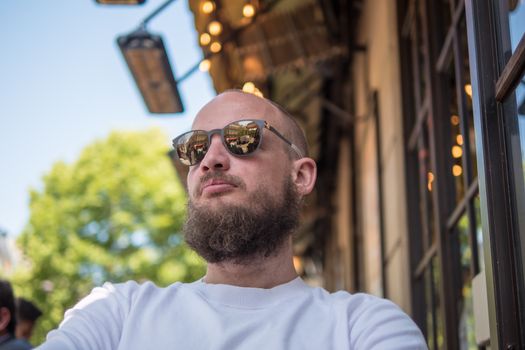 Handsome french bearded man with fashionable sunglasses and white shirt in Paris, sit in a coffee terrace