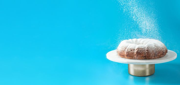 Icing sugar falling of fresh home made bundt cake. Powder sugar falls on fresh bunt cake over blue background. Copy space for text. Ideas and recipes for breakfast or dessert. Banner