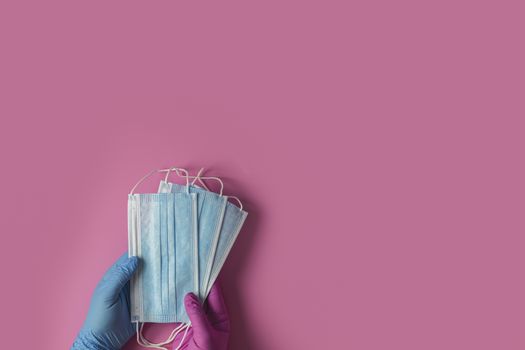 Doctor holds respiratory surgical face mask in hands blue and pink medical gloves on pink background. Pandemic corona virus, insurance, airborne diseases, SARS, grippe. 
