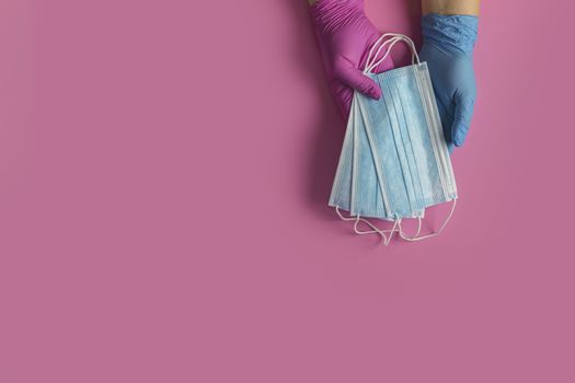 Doctor holds respiratory surgical face mask in hands blue and pink medical gloves on pink background. Pandemic corona virus, insurance, airborne diseases, SARS, grippe. 