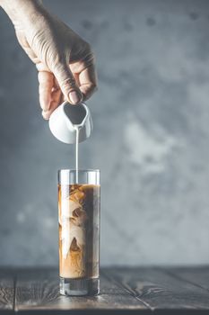 Women hand is pouring homemade sour cream from small jar to glass with brew cold coffee and ice. Cold summer drink on a dark wooden table and gray background with copy space.