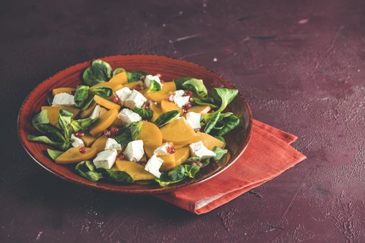 Healthy salad with persimmon, doucette (lambs-lettuce, cornsalad, feld salad) and feta cheese on a red plate on a red background. Superfoods Vitamin autumn or winter persimmon salad.