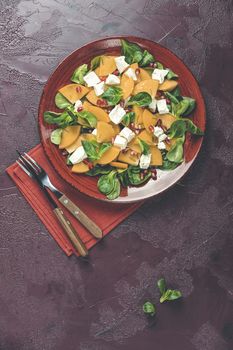 Healthy salad with persimmon, doucette (lambs-lettuce, cornsalad, feld salad) and feta cheese. Red surface. Superfoods Vitamin autumn or winter persimmon salad. Top view, flat lay, copy space.