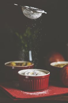 Sifting powdered sugar by sieve over apple pie in ceramic baking molds ramekin on dark wooden table. Close up, shallow depth of the field.