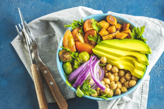 Healthy food. Healthy vegan lunch bowl of fried sweet potato, chickpeas, red onion, avocado, tomatoes, and brussels sprouts. Top view, classic blue concrete surface, flat lay.