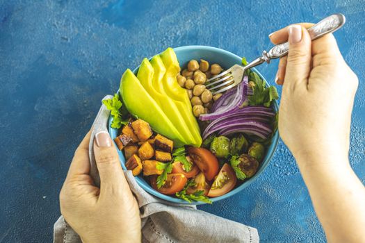 Woman is eating healthy lunch. Vegan lunch bowl of fried sweet potato, chickpeas, red onion, avocado, tomatoes, and brussels sprouts. Top view, flat lay. Clean and balanced healthy food concept