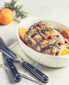 Roasted pork in white dish, christmas baked ham with cranberries, tangerines, thyme, rosemary, garlic on light table surface	