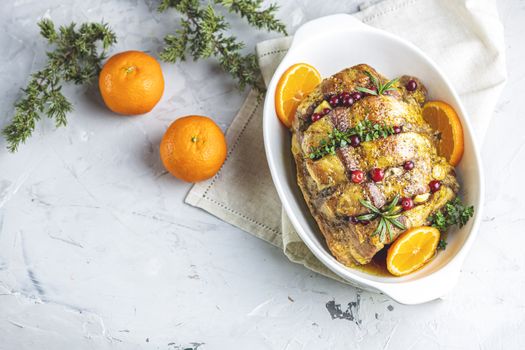 Roasted pork in white dish, christmas baked ham with cranberries, tangerines, thyme, rosemary, garlic on light table surface, close up.