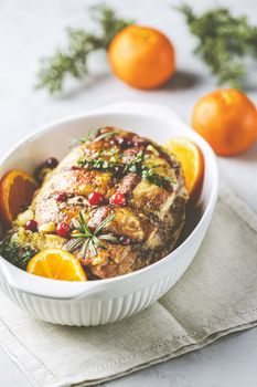 Roasted pork in white dish, christmas baked ham with cranberries, tangerines, thyme, rosemary, garlic on light table surface, close up.