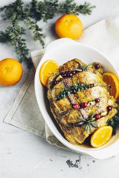 Roasted pork in white dish, christmas baked ham with cranberries, tangerines, thyme, rosemary, garlic on light table surface	