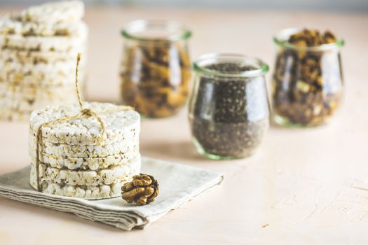American puffed rice cakes. Healthy snacks with almonds, raisins, peanuts, pistachios in glass jars on light pink concrete surface.	