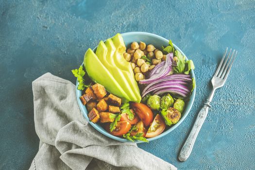 Healthy food. Healthy vegan lunch bowl of fried sweet potato, chickpeas, red onion, avocado, tomatoes, and brussels sprouts. Top view, classic blue concrete surface, flat lay