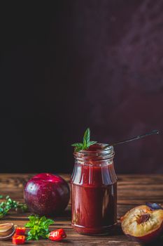Homemade DIY natural canned hot plum sauce chutney with chilli or tkemali in glass jar standing on wooden table with ingredients — plum, salt, pepper,  herbs, selective focus