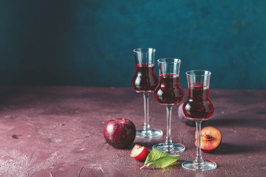 Plums strong alcoholic drink in grappas wineglass with dew. Hard liquor, slivovica, plum brandy or plum vodka with ripe plums on dark blue and  claret bordeaux concrete surface.