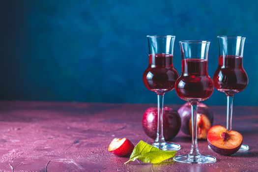 Plums strong alcoholic drink in grappas wineglass with dew. Hard liquor, slivovica, plum brandy or plum vodka with ripe plums on dark blue and claret bordeaux concrete surface