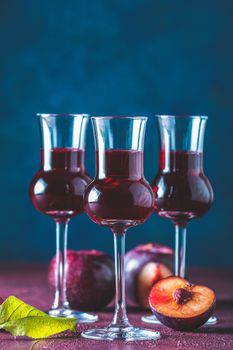 Plums strong alcoholic drink in grappas wineglass with dew. Hard liquor, slivovica, plum brandy or plum vodka with ripe plums on dark blue and  claret bordeaux concrete surface.