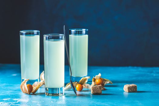 Alcoholic cocktail with Ouzo greek anise brandy or Arabic alcohol drink Raki in highball glass, decorated Physalis peruviana and brown sugar on blue concrete surface.