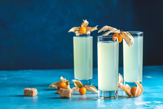 Alcoholic cocktail with Ouzo greek anise brandy or Arabic alcohol drink Raki in highball glass, decorated Physalis peruviana and brown sugar on blue concrete surface.