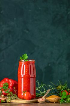 Homemade DIY natural canned hot tomato sauce chutney with chilli or adjika in glass jar standing on wooden table with cherry tomatoes, salt, pepper,  herbs, selective focus