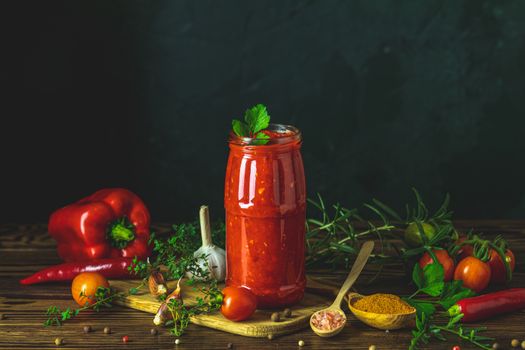 Homemade DIY natural canned hot tomato sauce chutney with chilli or adjika in glass jar standing on wooden table with cherry tomatoes, salt, pepper, herbs, selective focus