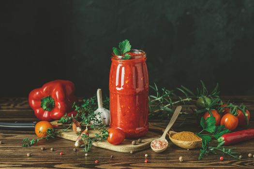 Homemade DIY natural canned hot tomato sauce chutney with chilli or adjika in glass jar standing on wooden table with cherry tomatoes, salt, pepper, herbs, selective focus