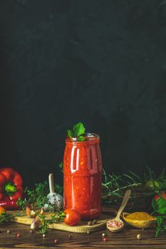Homemade DIY natural canned hot tomato sauce chutney with chilli or adjika in glass jar standing on wooden table with cherry tomatoes, salt, pepper,  herbs, selective focus