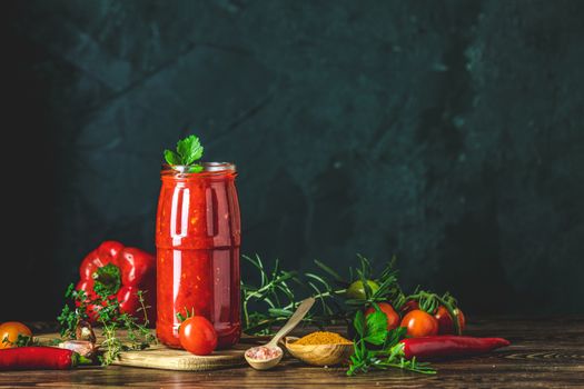 Homemade DIY natural canned hot tomato sauce chutney with chilli or adjika in glass jar standing on wooden table with cherry tomatoes, salt, pepper,  herbs, selective focus
