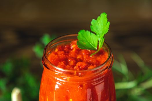 Homemade DIY natural canned hot tomato sauce chutney with chilli or adjika in glass jar standing on wooden table with cherry tomatoes, salt, pepper,  herbs, selective focus