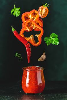 Sauce components frozen in the air. Homemade DIY natural canned hot tomato sauce chutney with chilli or adjika in glass jar standing on wooden table with flying ingredients, selective focus