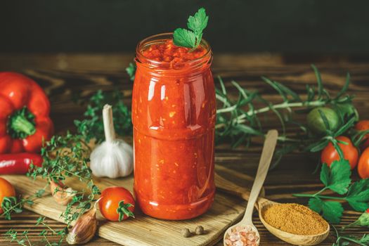 Homemade DIY natural canned hot tomato sauce chutney with chilli or adjika in glass jar standing on wooden table with cherry tomatoes, salt, pepper,  herbs, selective focus