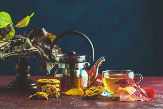 Autumn teatime composition on dark background with colored leaves and chocolate cookies, sun light beam on the cup behind the glass with water dew drops, selective focus