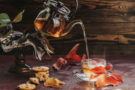 Pour hot tea in glass cup. Autumn teatime composition on dark background with colored leaves and chocolate cookies, sun light beam on the cup.