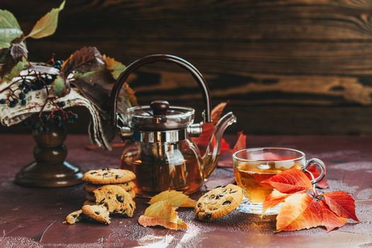 Autumn teatime composition on dark background with colored leaves and chocolate cookies, sun light beam on the cup behind the glass with water dew drops, selective focus