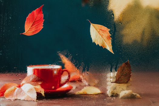 Autumn teatime blurred composition with colored leaves on glass with water dew drops. Red in white dotted cup of hot tea behind the glass out of focus