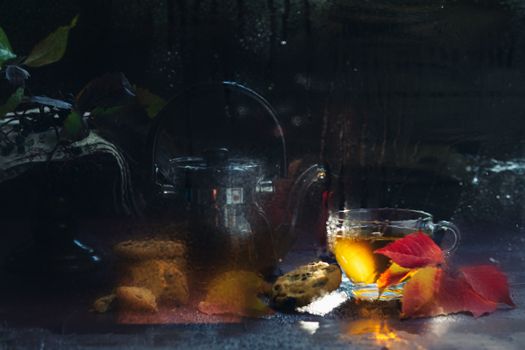 Autumn teatime blurred composition on dark background with colored leaves, sun light beam on the cup behind the glass with water dew drops, out of focus.