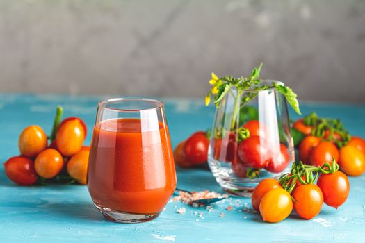 Glasses of fresh delicious jummy red tomato juice and fresh raw tomatoes with pink salt in spoon on light concrete surface. Close up. Gmo free. Natural good food.