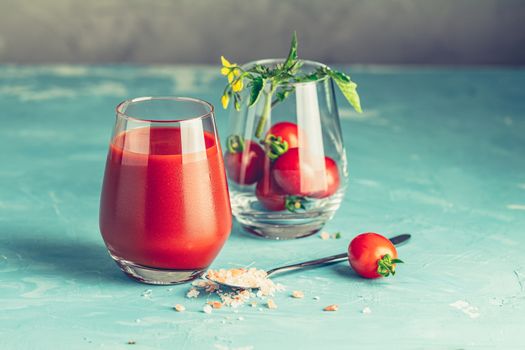 Glasses of fresh delicious jummy red tomato juice and fresh raw tomatoes with pink salt in spoon on light concrete surface. Close up. Gmo free. Natural good food.